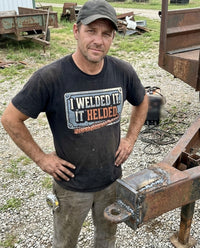 Load image into Gallery viewer, Welder wearing the I Welded It It Helded shirt next to a trailer hitch he welded
