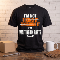 Load image into Gallery viewer, I'm Not Fixing It, I'm Waiting On Parts mechanic t-shirt standing on a workbench with a parts box, wrench, mug and work gloves