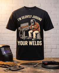 Load image into Gallery viewer, I'm Silently Judging Your Welds welder t-shirt on a workbench with a welding helmet, gloves, C-clamp and MIG gun